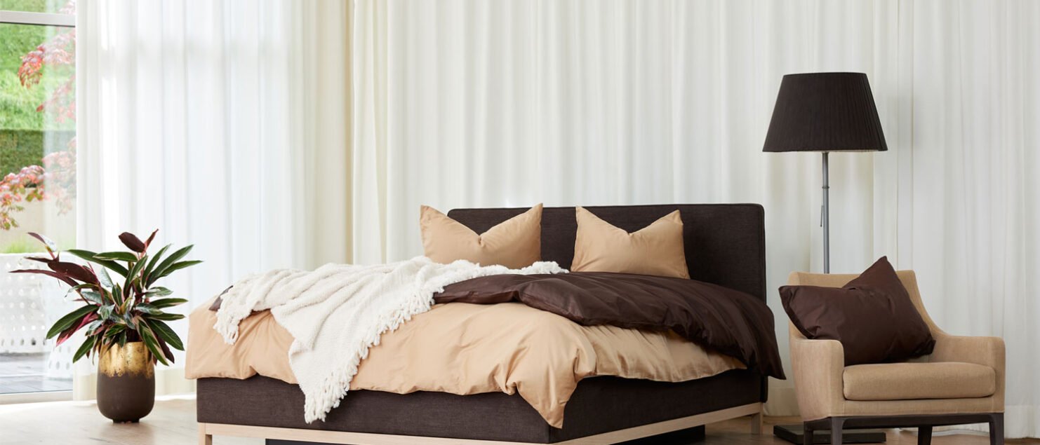 Ein braunes Bett mit beigen Duvet und Kissen steht einem schön dekorierten Zimmer mit langen weissen Vorhängen.