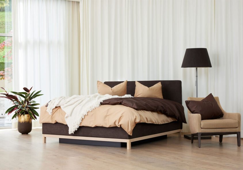 Ein braunes Bett mit beigen Duvet und Kissen steht einem schön dekorierten Zimmer mit langen weissen Vorhängen.